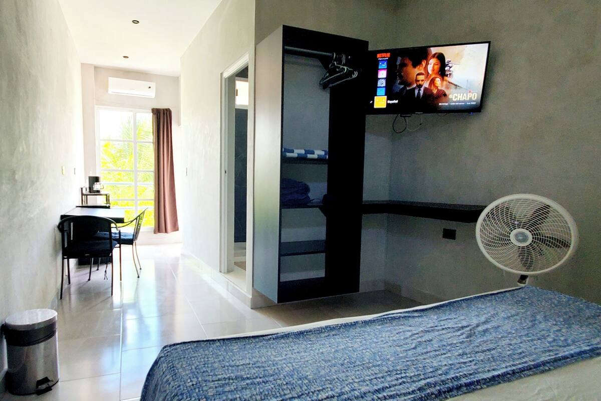 Ein Hotelzimmer mit Bett, einem an der Wand montierten Fernseher, einem Schreibtisch mit Stuhl und einem Ventilator.