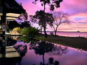 A waterfront view with a reflection in the water, trees, and a building with outdoor seating.