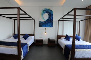 Una habitación con dos camas, cada una con estructura de madera y ropa de cama azul. Hay una lámpara de pared y un cuadro de una ola.