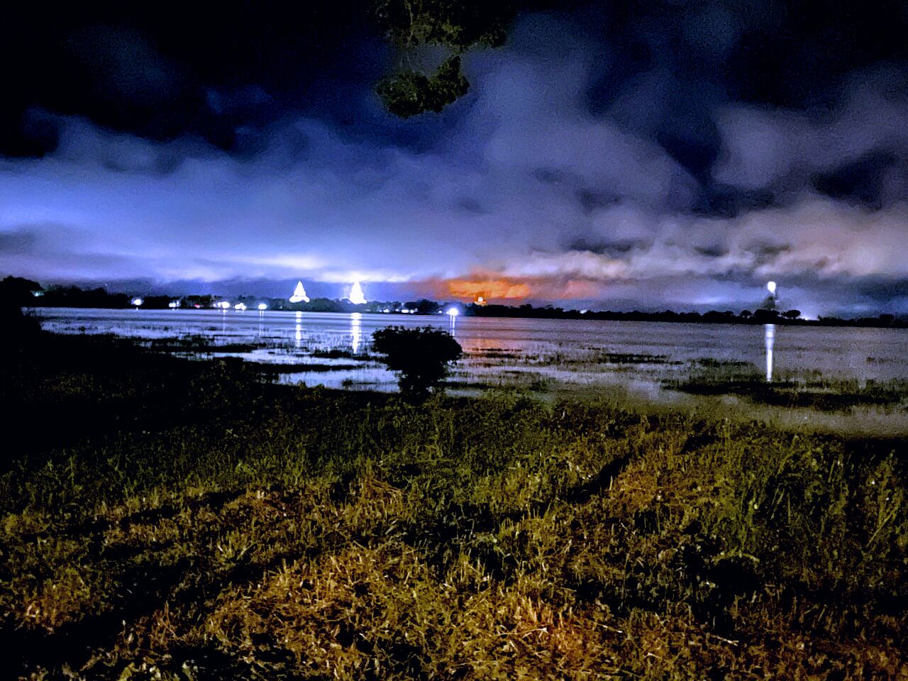 Un paysage nocturne avec une étendue d’eau, un premier plan herbeux et une vue lointaine sur les contours d’une ville, avec des bâtiments illuminés.