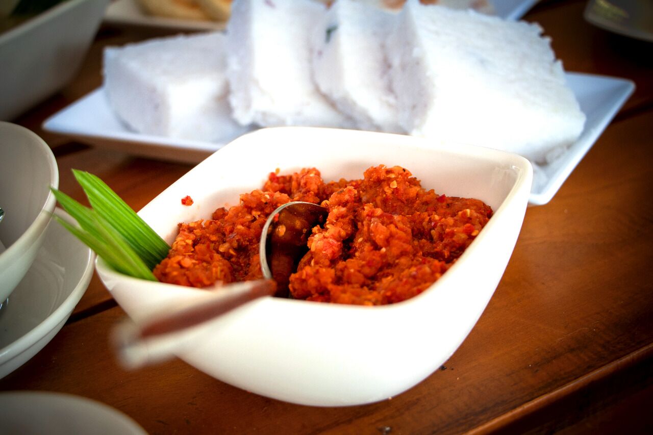 A white bowl filled with a red dish, possibly a curry, with a spoon in it. In the background, there is a plate with white rice and a white bowl with green leaves.