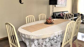 Un comedor con una mesa redonda, sillas blancas, mantel de encaje, una cafetera y una lámpara de pared.