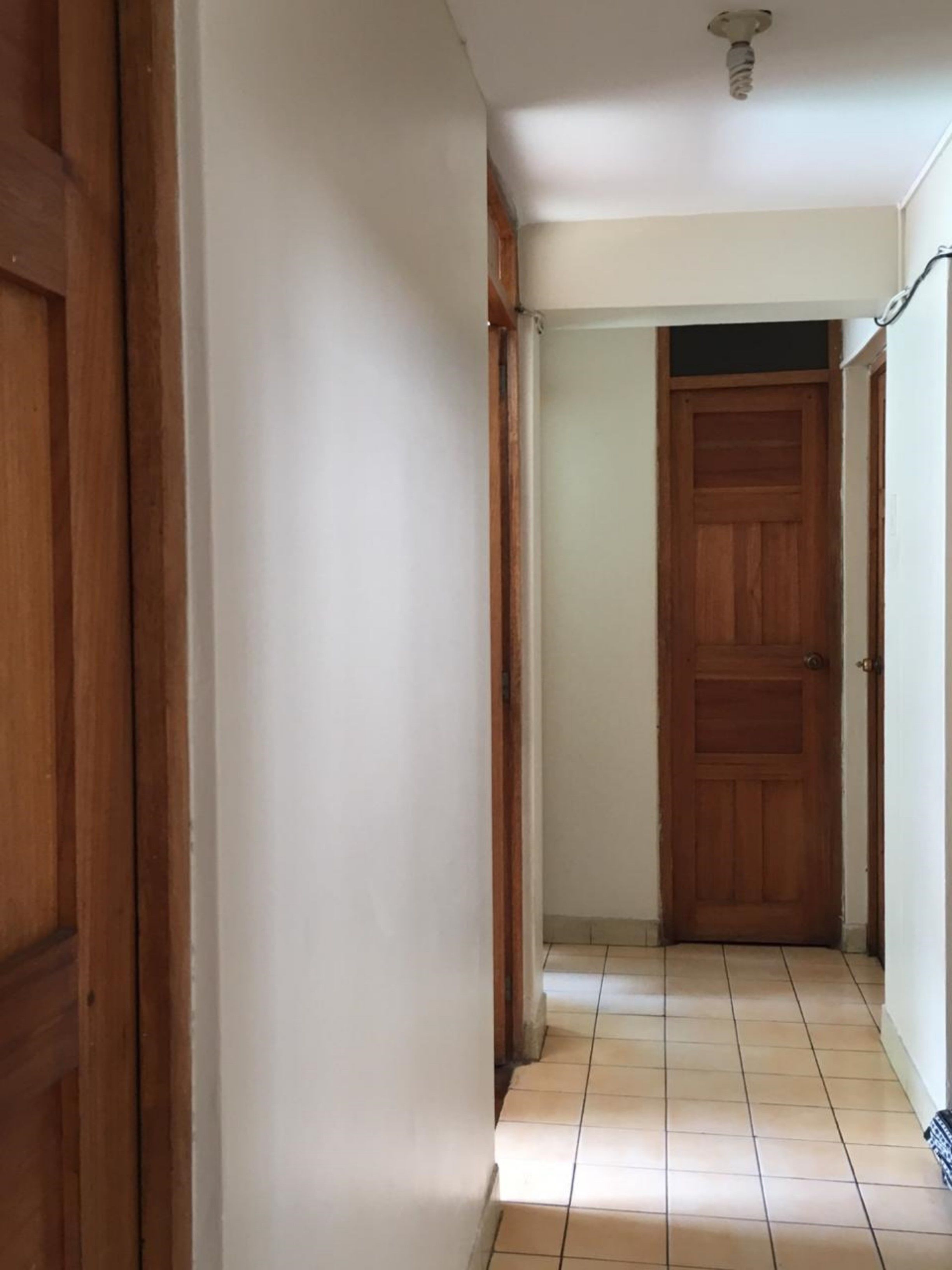 Un pasillo con una puerta de madera al final, paredes blancas y suelo de baldosas.