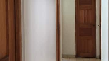Un pasillo con una puerta de madera al final, paredes blancas y suelo de baldosas.