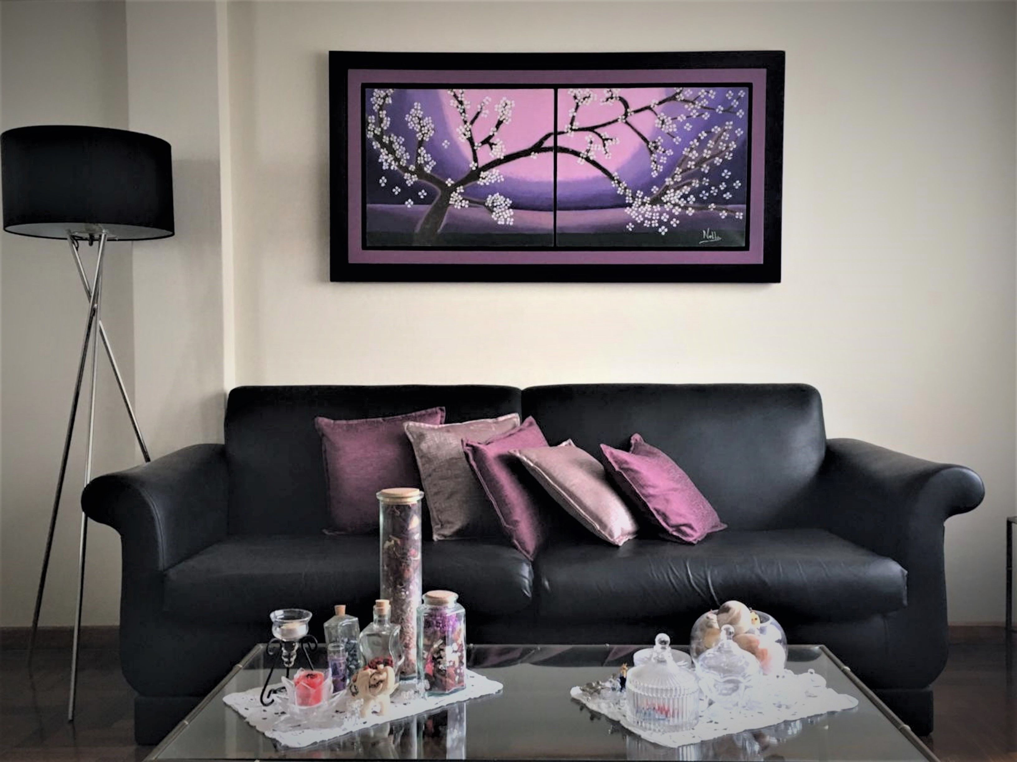 Un soggiorno moderno con un divano in pelle nera, cuscini viola, un tavolino da caffè in vetro e oggetti decorativi, e un quadro di un albero appeso al muro.