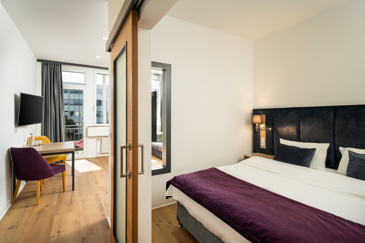 Quarto de hotel com cama, escrivaninha, cadeira, televisão e janela.