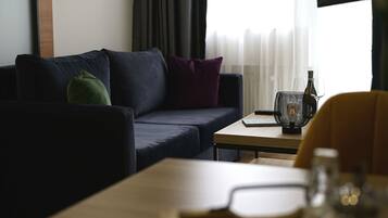 Una sala moderna con un sofá azul oscuro, cojines verdes y morados, una mesa de centro de madera y una mesita auxiliar con una vela y un vaso.