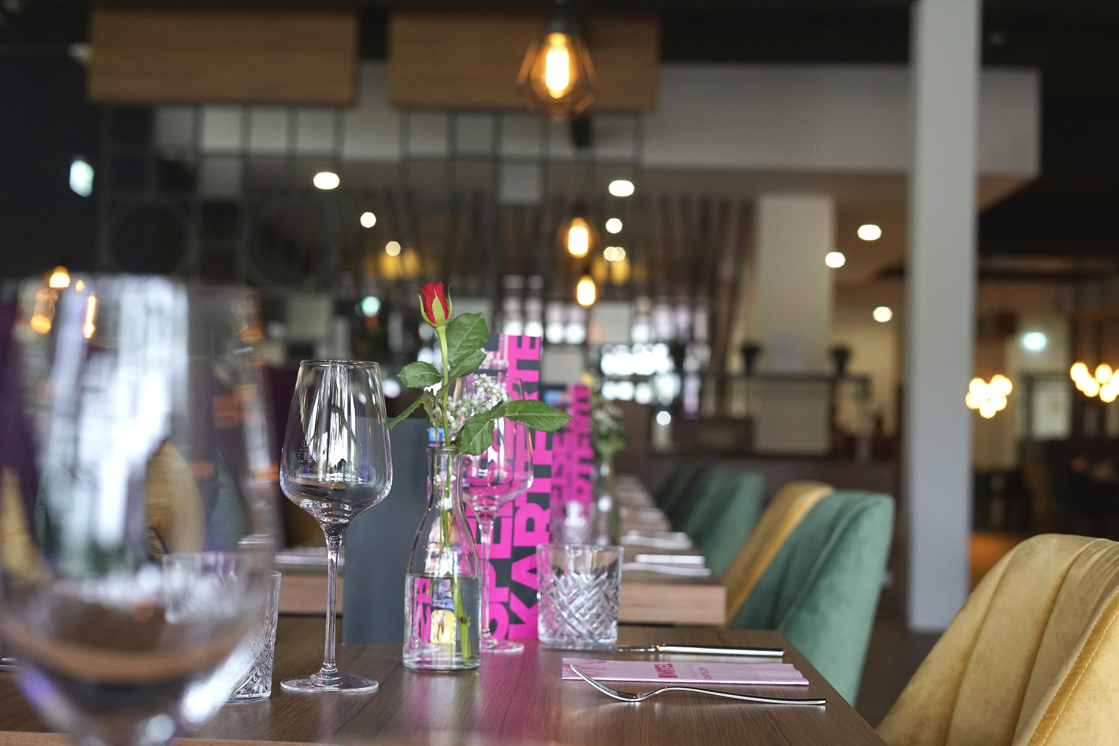 A restaurant interior with a long table set for dining, featuring clear glasses, a vase with a flower, and a menu.