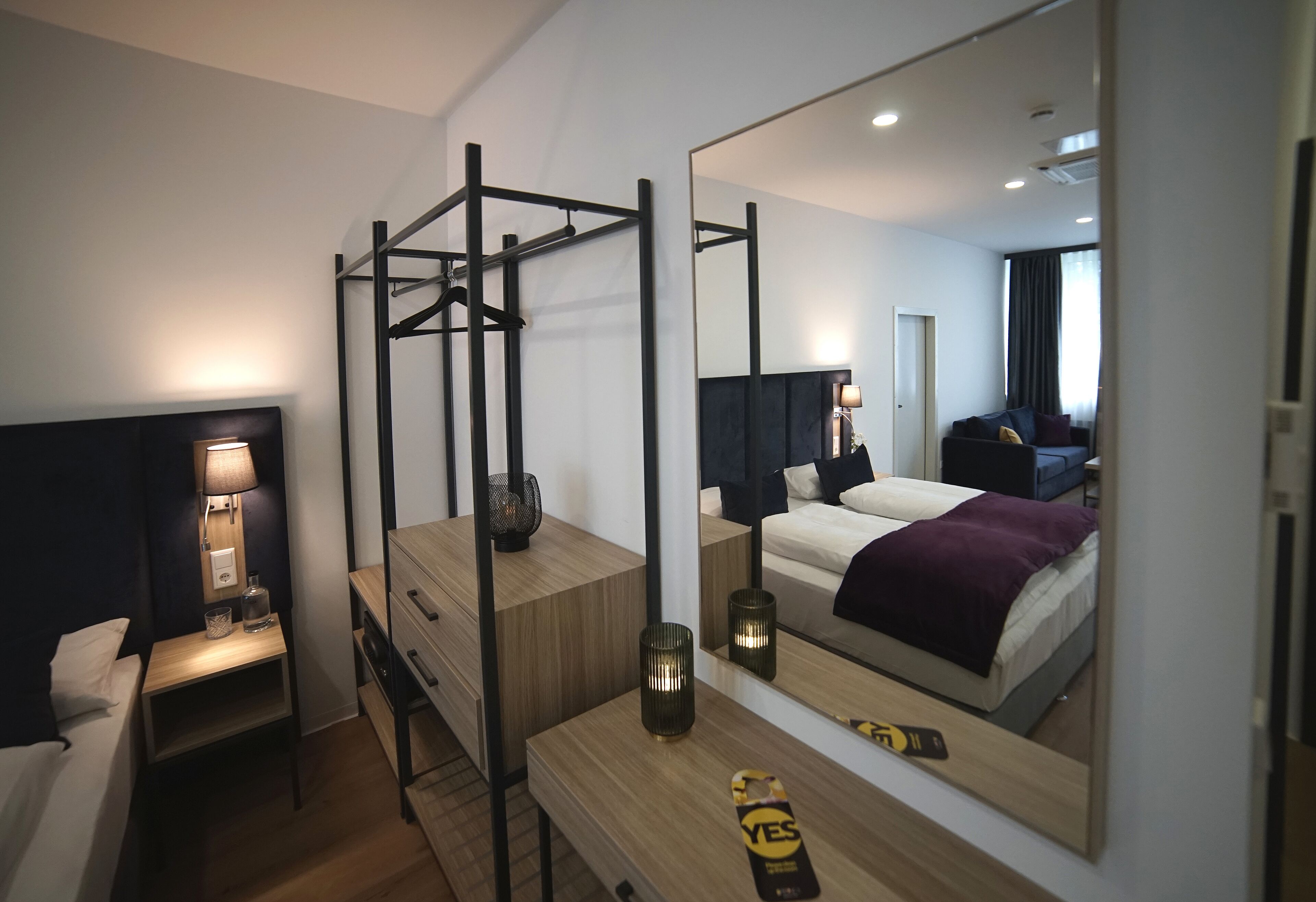 A modern hotel room with a bed, a desk, a chair, a mirror, and a hanging light fixture.