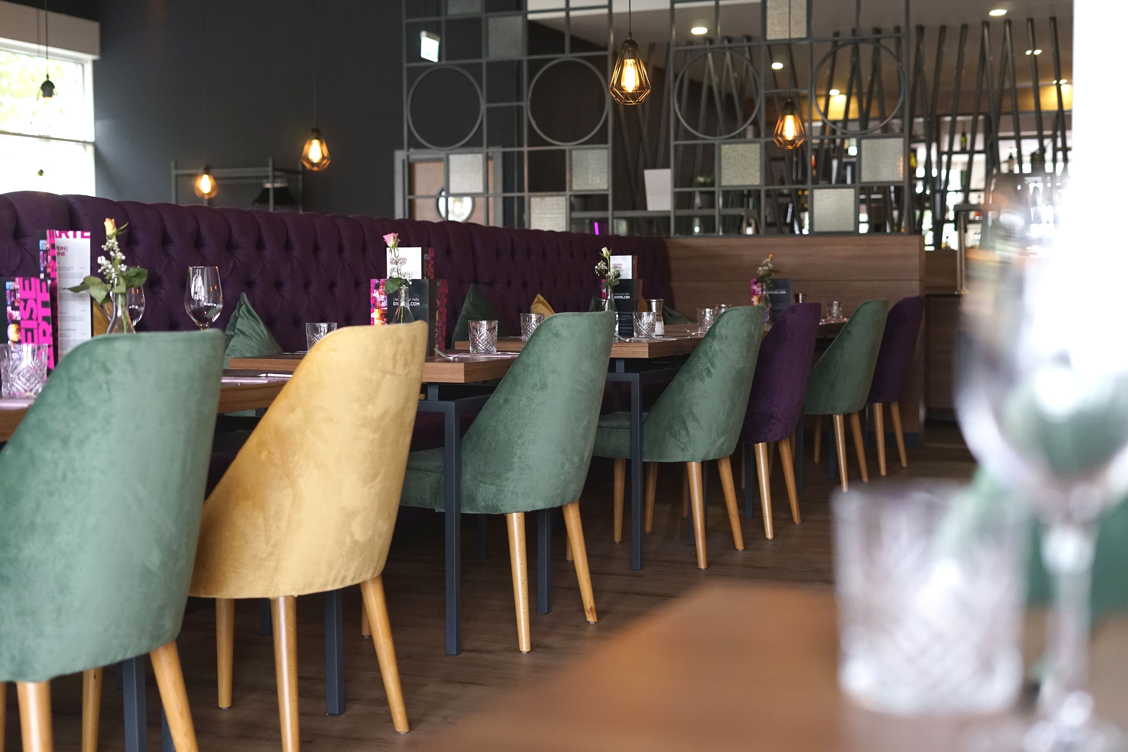 Interno di un ristorante dal design moderno, con sedie imbottite in verde e giallo, tavoli in legno e lampade a sospensione.
