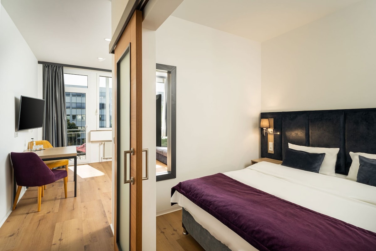 Habitación de hotel con cama, escritorio, silla, televisión y vista al exterior.