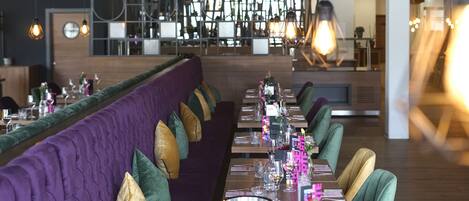 A modern restaurant with a long dining table, cushioned seating, and pendant lighting.