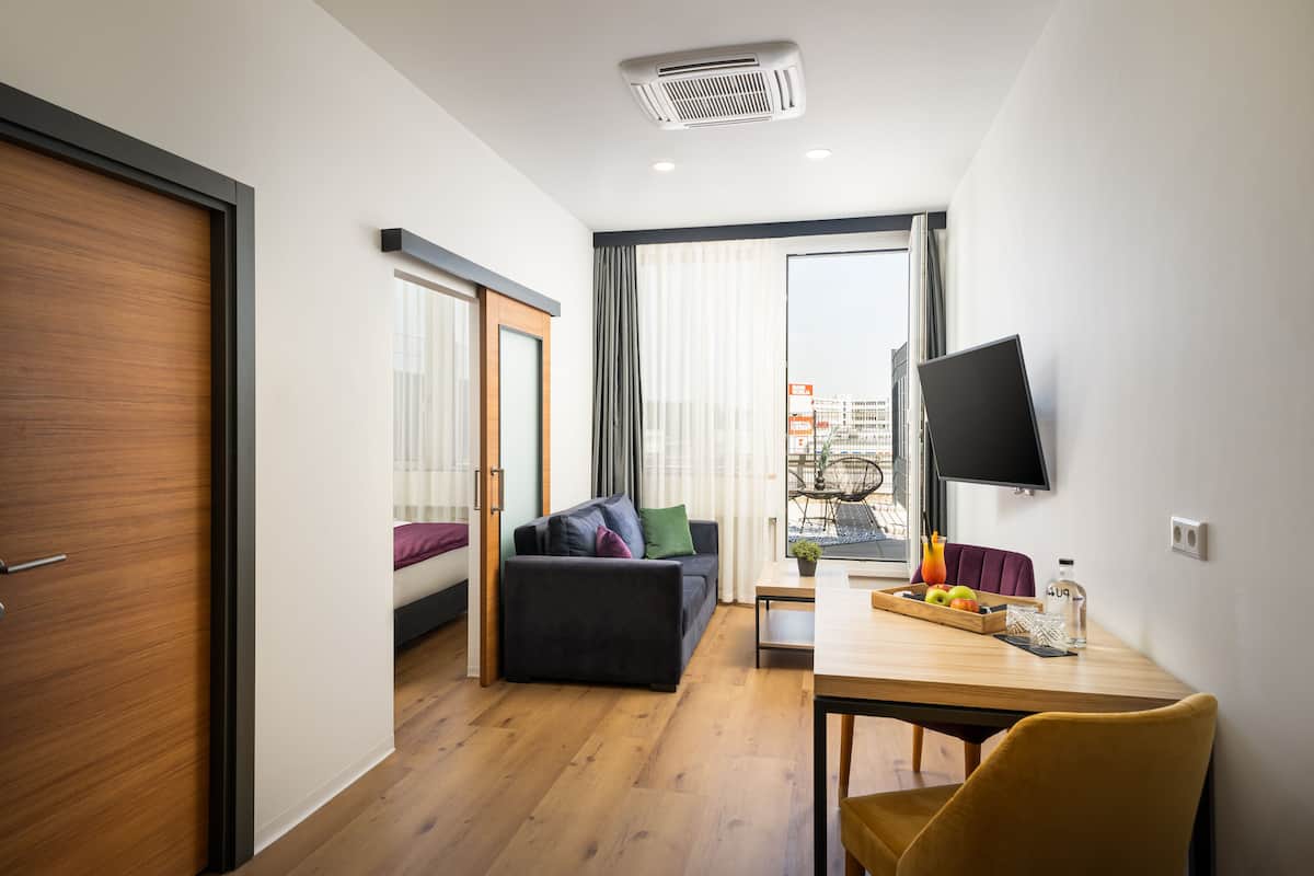 Une chambre d’hôtel moderne avec un plancher en bois, un canapé, une table à manger agrémentée d’un panier de fruits, un téléviseur fixé au mur et une porte coulissante.