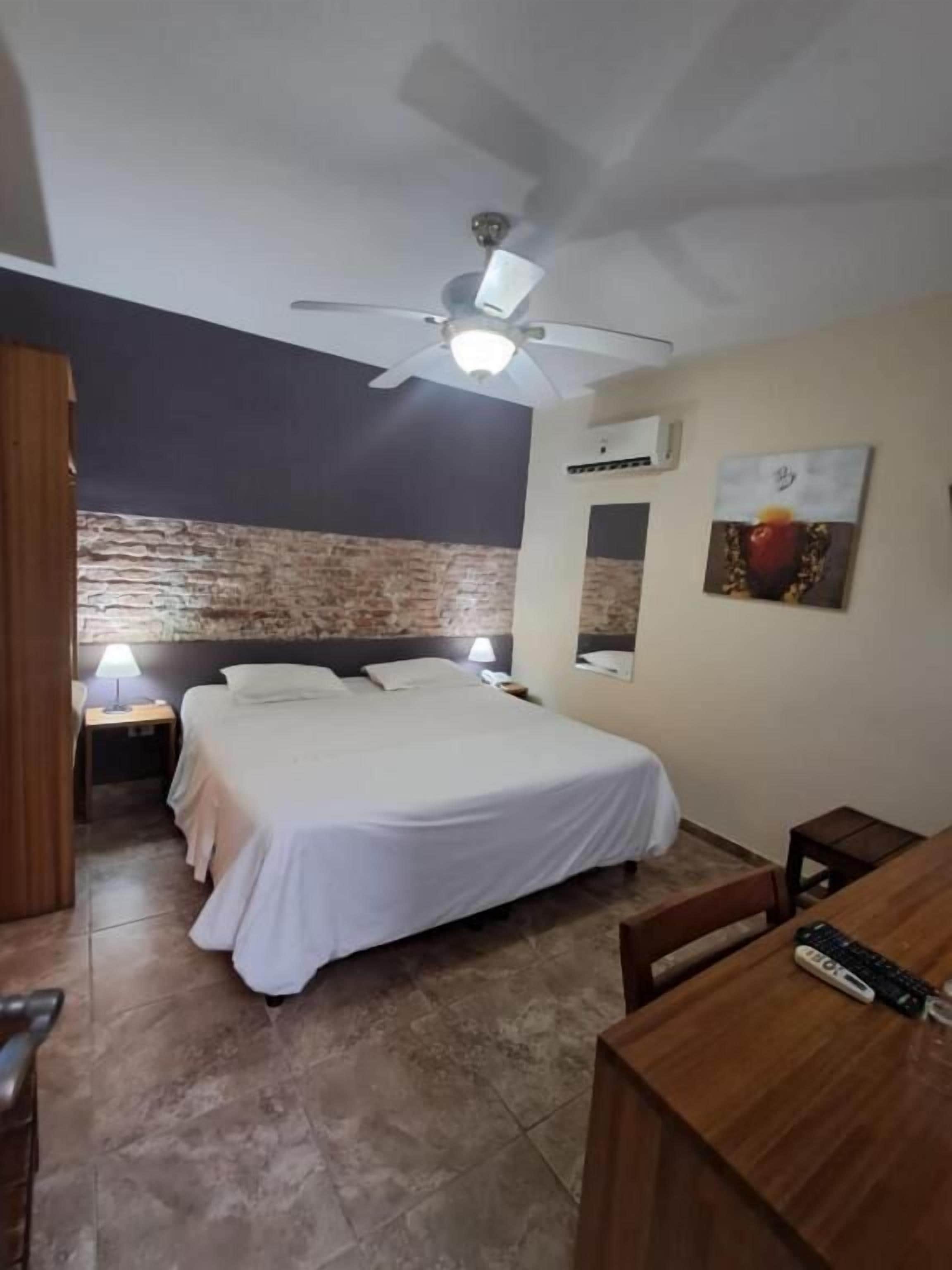 Ett hotellrum med en säng, ett skrivbord, en takfläkt, en väggmonterad luftkonditioneringsanläggning och en tavla.