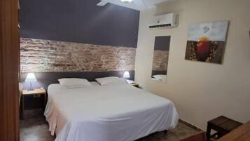 Ett hotellrum med en säng, ett skrivbord, en takfläkt, en väggmonterad luftkonditioneringsanläggning och en tavla.