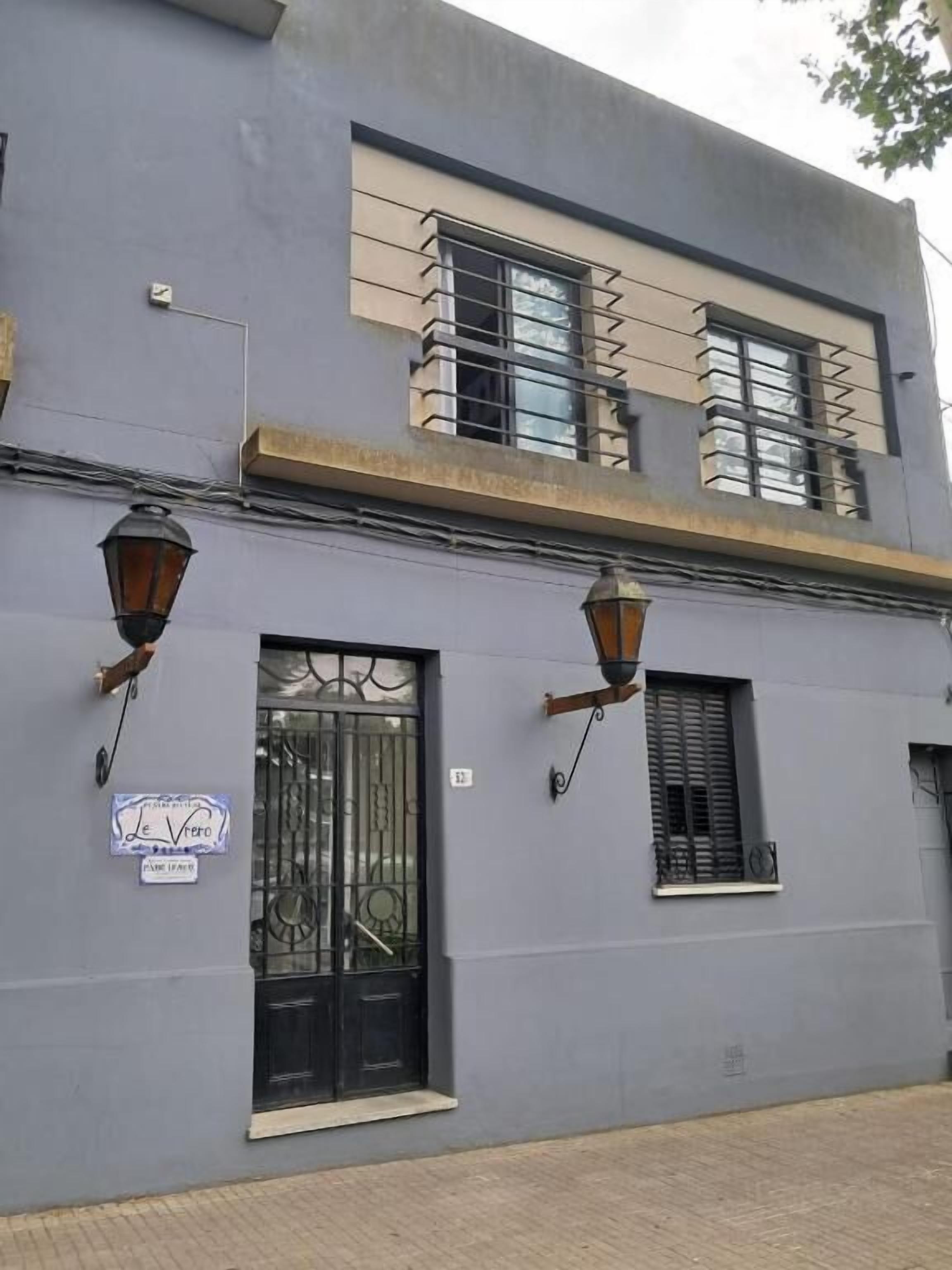 Un edificio de dos pisos con una reja de metal negro, un letrero encima de la reja y dos ventanas con contraventanas.