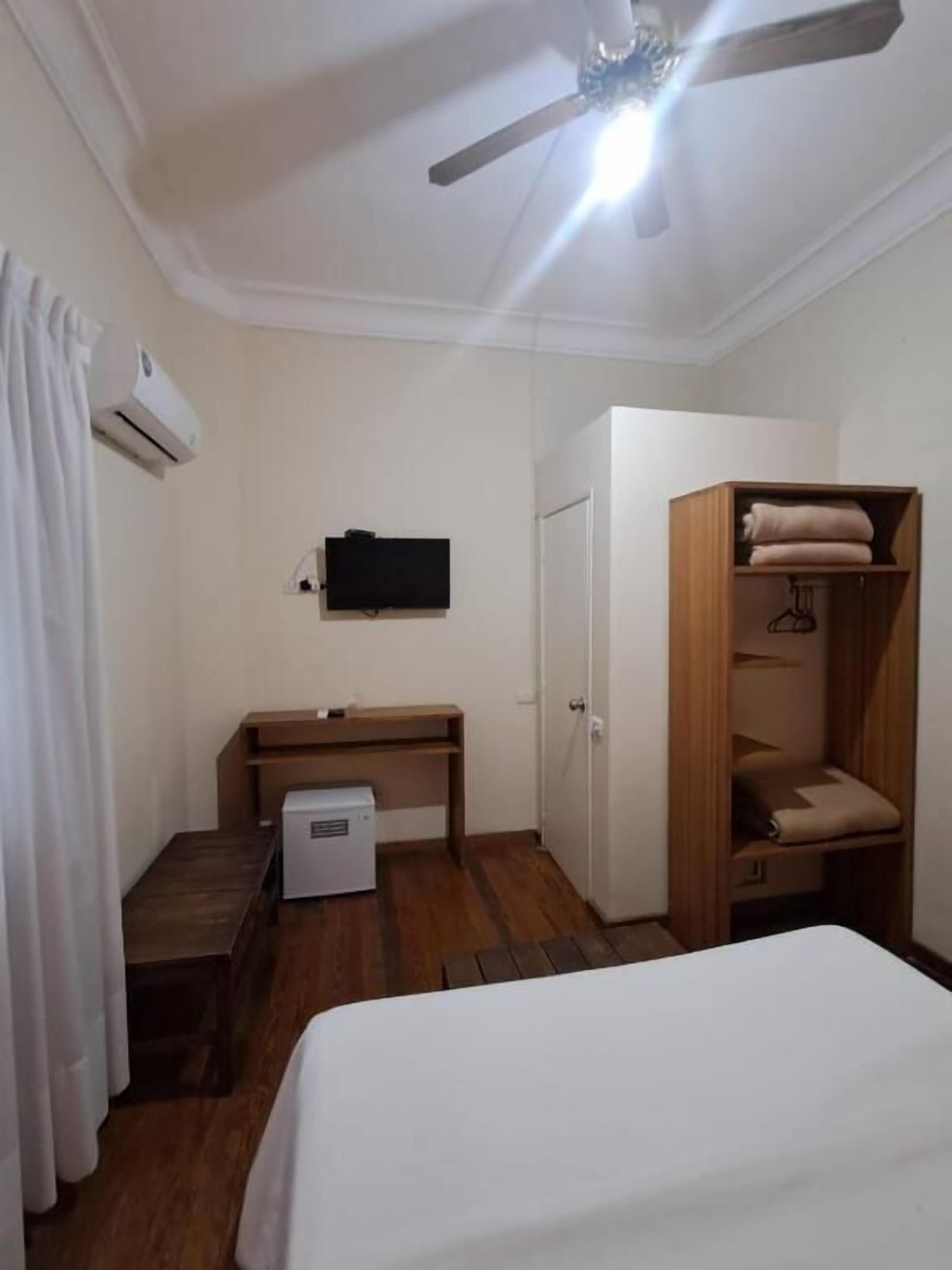 Ein kleines Hotelzimmer mit Bett, Schreibtisch, Kleiderschrank und Deckenventilator.