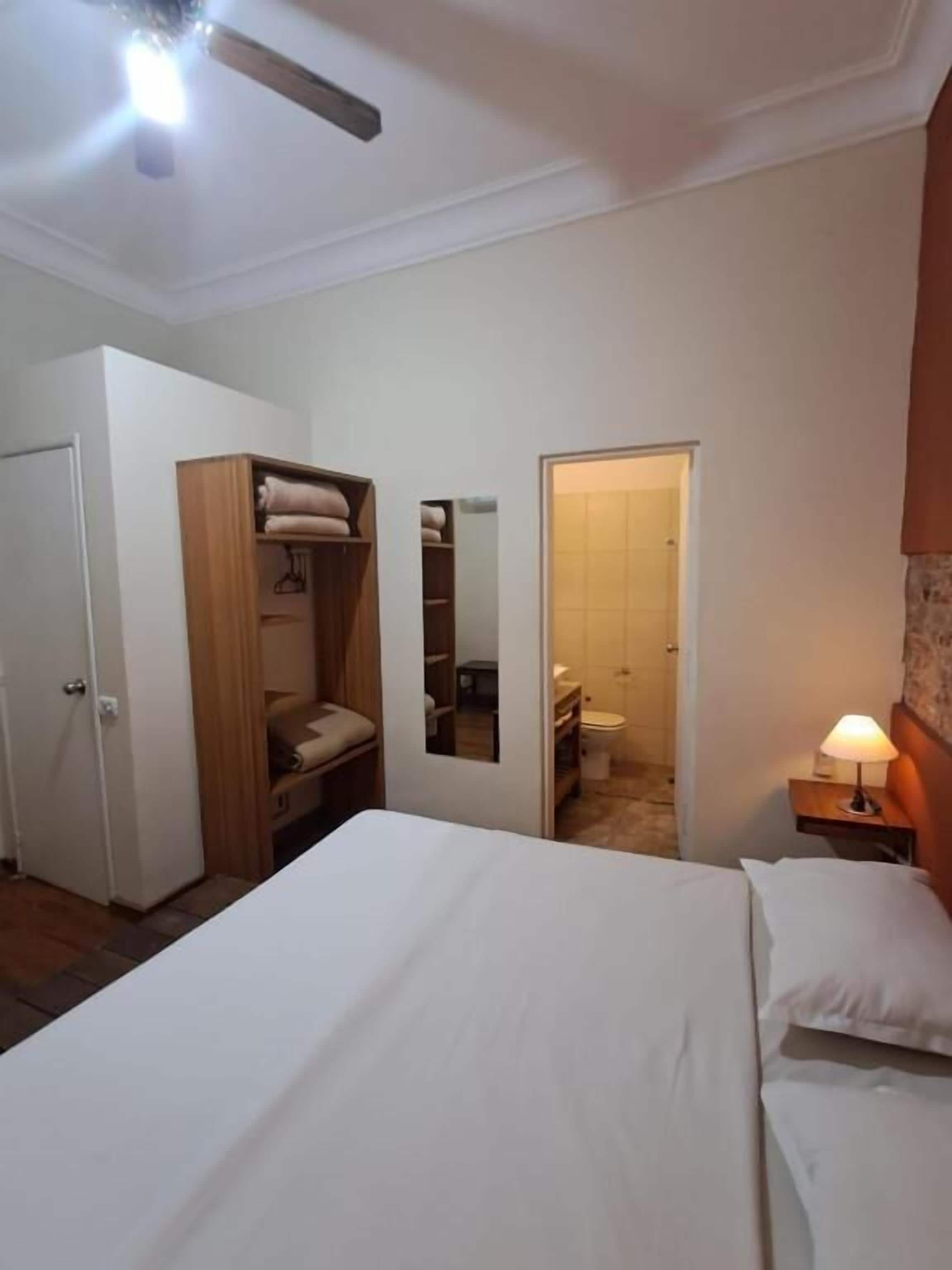 Un dormitorio con cama, armario, espejo, mesita de noche con lámpara y puerta que da a un baño.