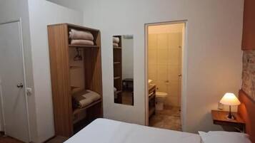 Un dormitorio con cama, armario, espejo, mesita de noche con lámpara y puerta que da a un baño.
