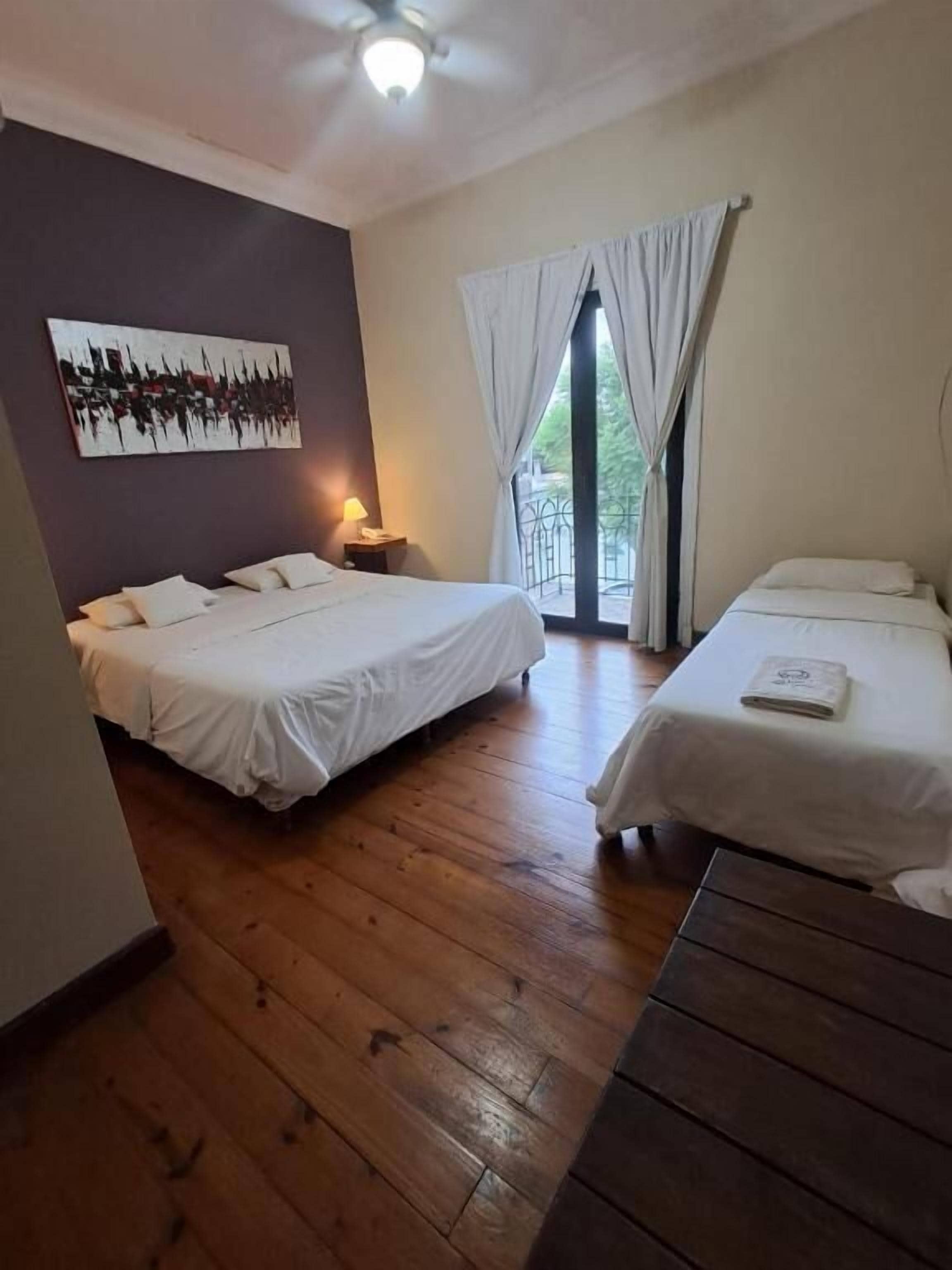 Ein Zimmer mit zwei Betten, Holzboden, Deckenventilator und einem Fenster mit Vorhängen.