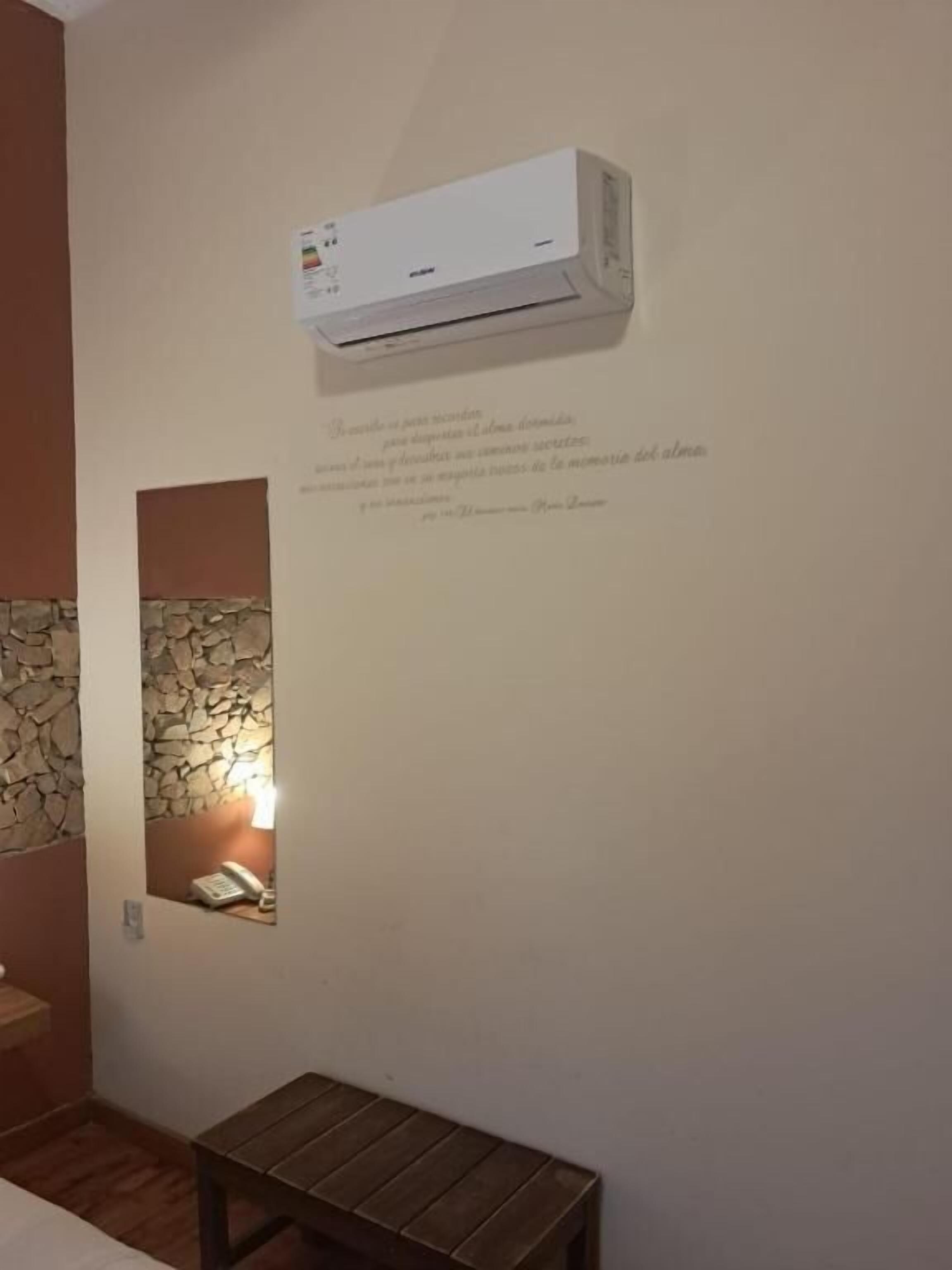 Un aire acondicionado de pared, un banco pequeño, y una pared con textura y un accesorio de iluminación.