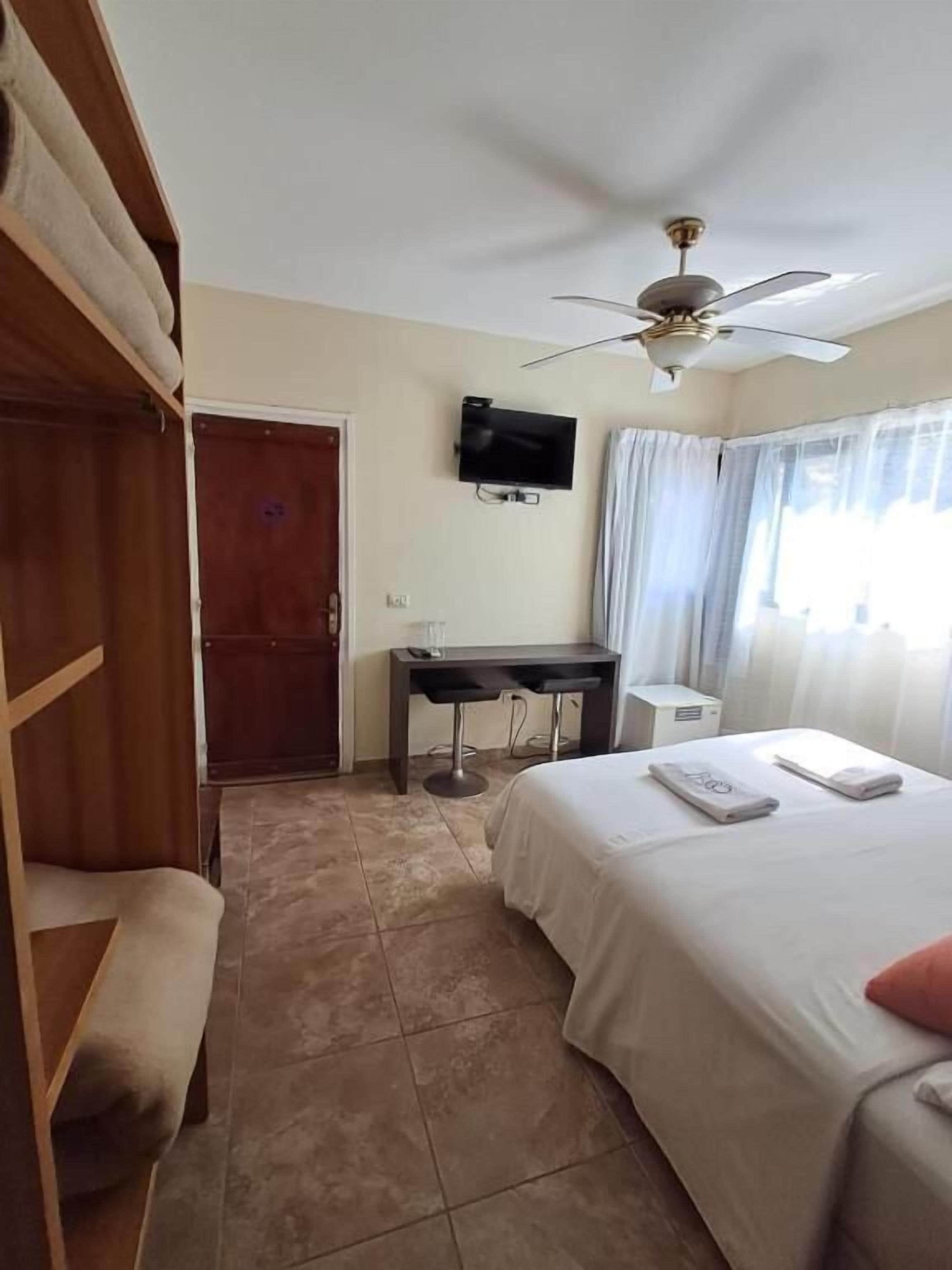 A hotel room with a bed, a desk, a chair, a wardrobe, a ceiling fan, and a television.