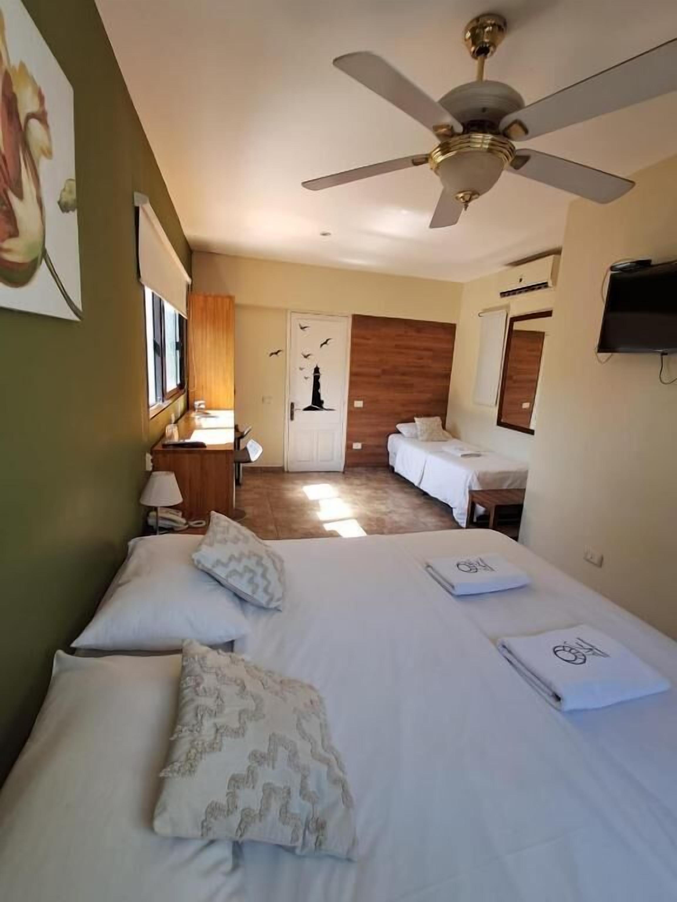 Quarto de hotel com cama, ventoinha no teto, televisão e porta de acesso a outro espaço.