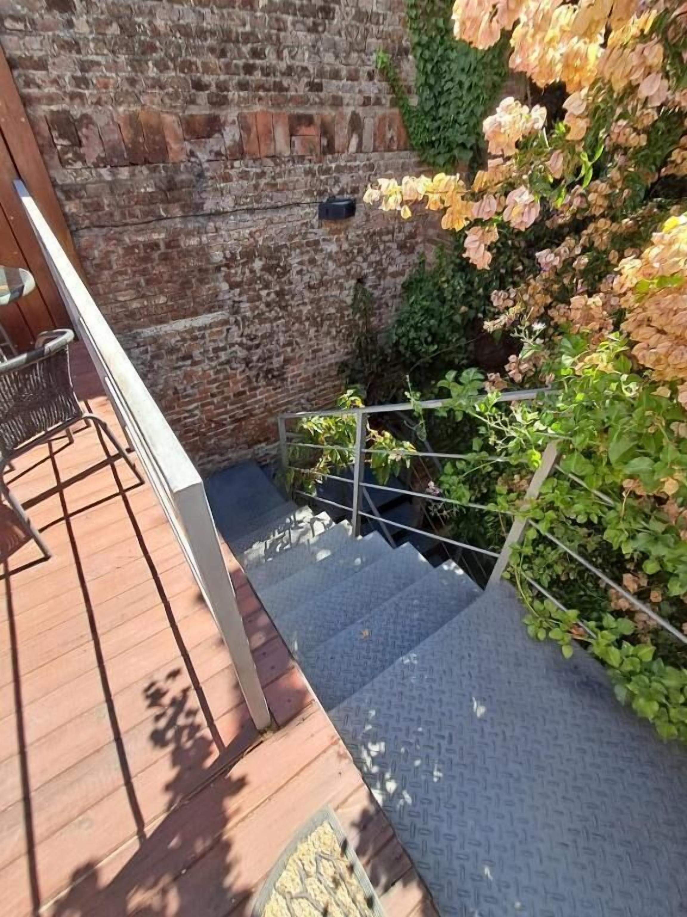 Eine Holzterrasse mit Metallgeländer, eine Backsteinmauer und eine Treppe, die zu einer niedrigeren Ebene führt.