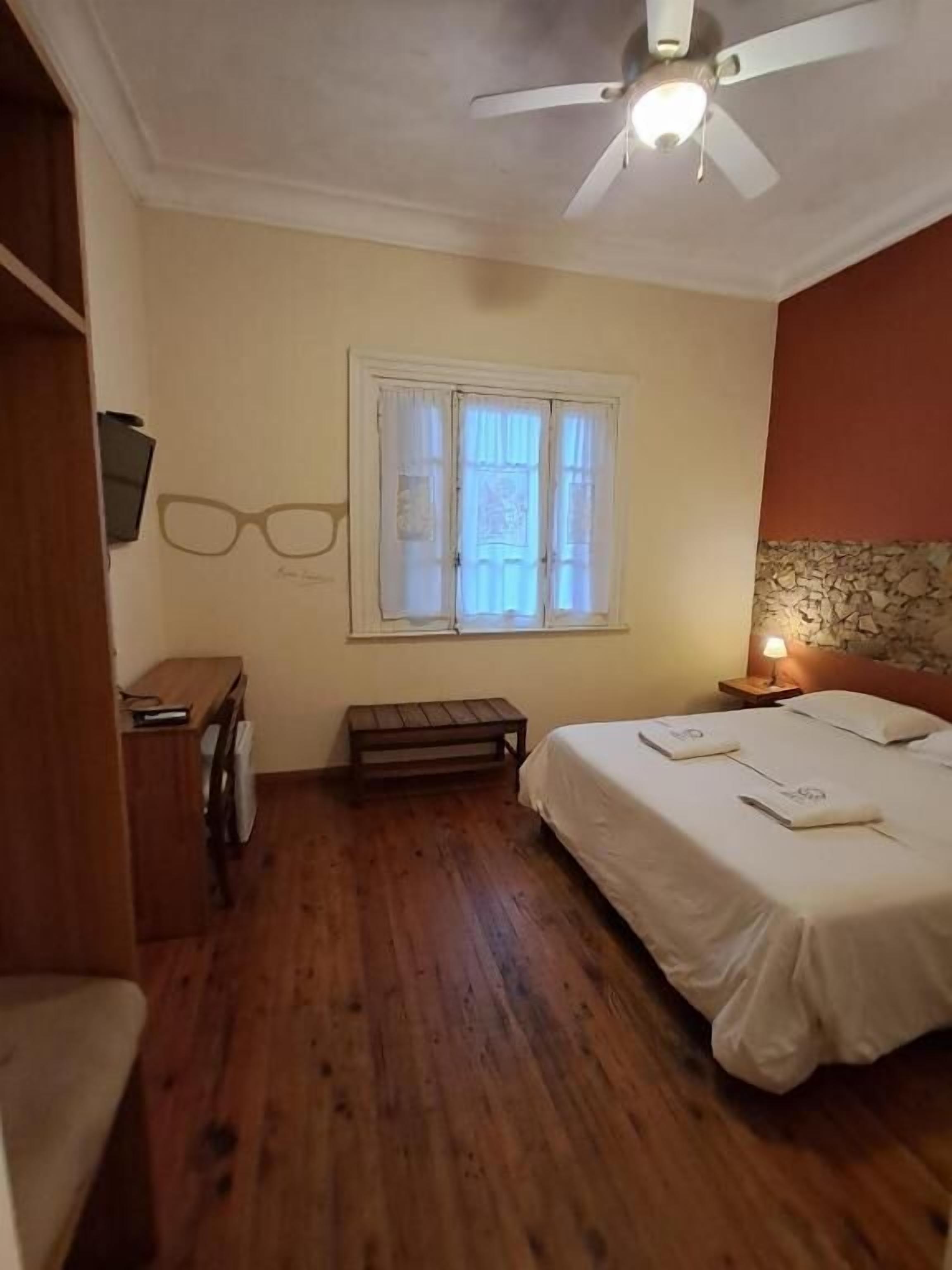 Un dormitori amb un llit, escriptori, cadira, finestra amb cortines, ventilador de sostre i una meseta de nit amb làmpada.