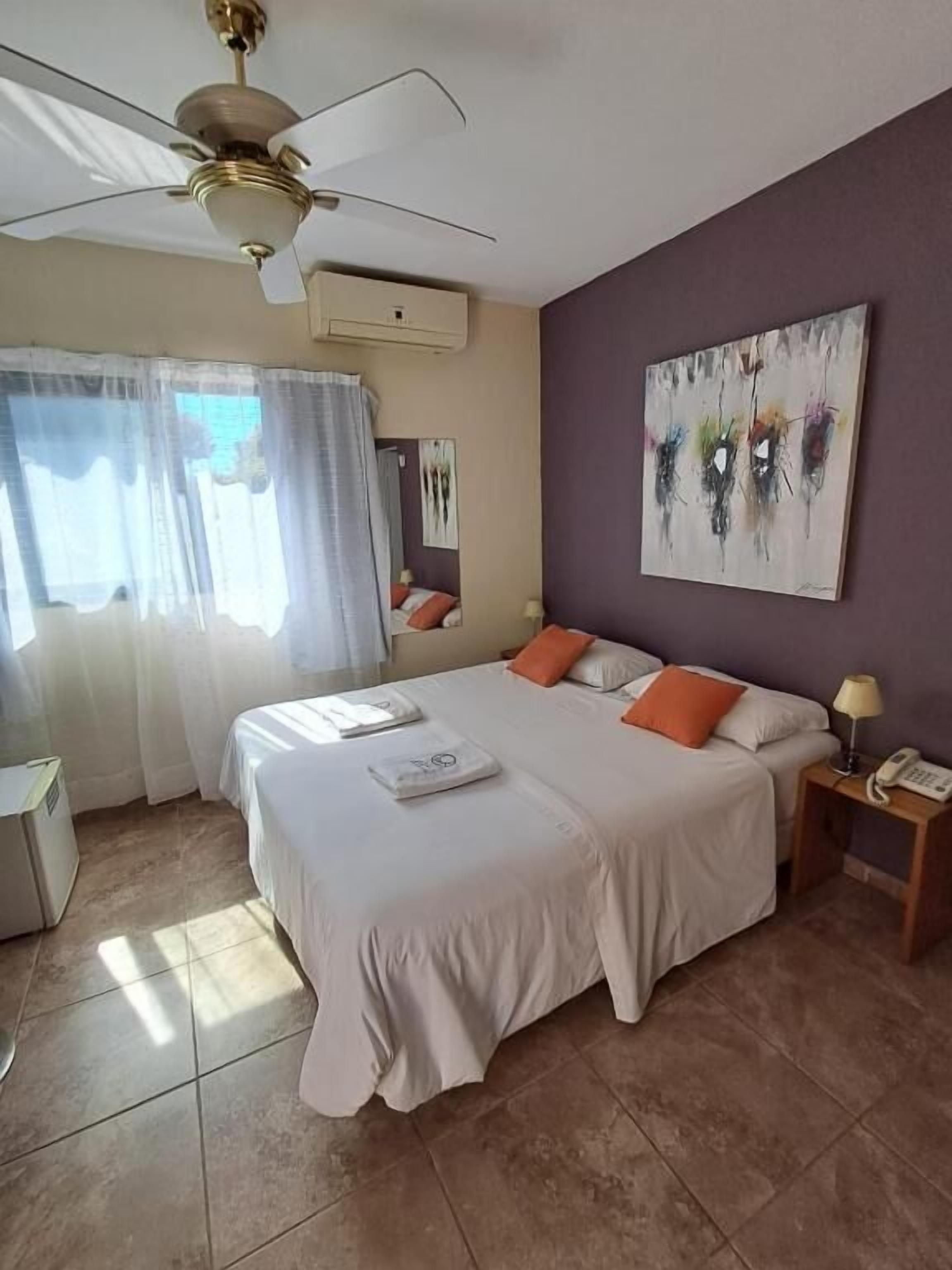 Habitación de hotel con cama, ventilador de techo, un cuadro en la pared y una mesita de noche con lámpara.