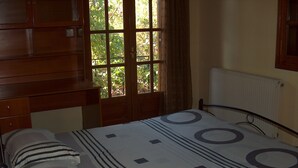 A bedroom with a bed, a nightstand, a lamp, a window with curtains, and a radiator. - Red Sun (Chios)