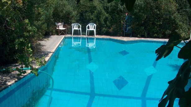 A swimming pool with a tiled edge, surrounded by greenery and two white plastic chairs. - Red Sun (Chios)