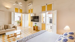 Une chambre d’hôtel avec un lit, un canapé, une table basse, une télévision et une fenêtre avec des volets jaunes.