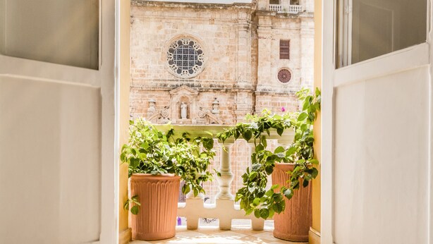 Une porte ouverte donne sur une cour intérieure dotée d’un sol à carreaux, de plantes en pot et offrant une vue sur un bâtiment historique surmonté d’une tour horloge.