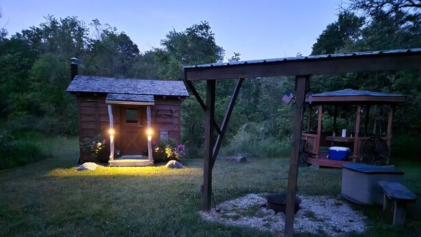 A rustic cabin with a porch illuminated by lights, a wooden deck, and a small outdoor seating area. - Unplug at cabin for 2 (Polk)