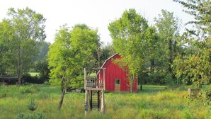 A red barn with a wooden platform and a ladder leading up to it, surrounded by lush greenery and trees. - Unplug at cabin for 2 (Polk)