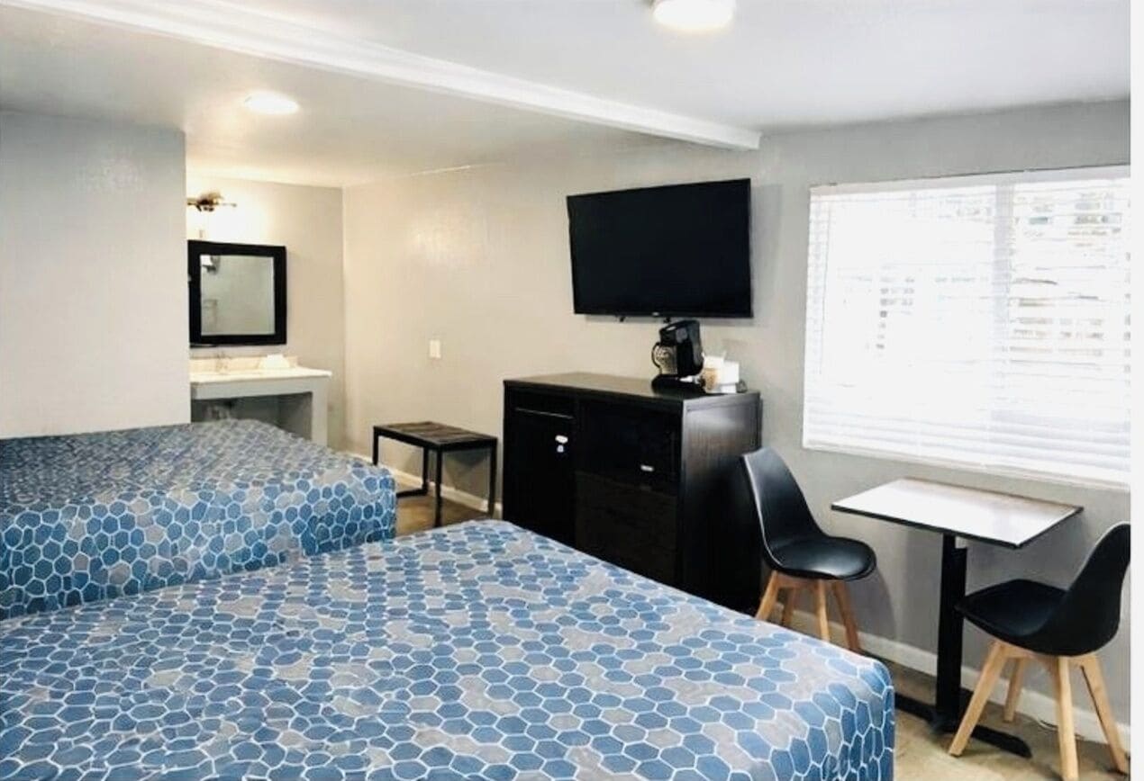 Une chambre avec deux lits, un bureau avec des chaises, une télévision et une fenêtre avec des stores.