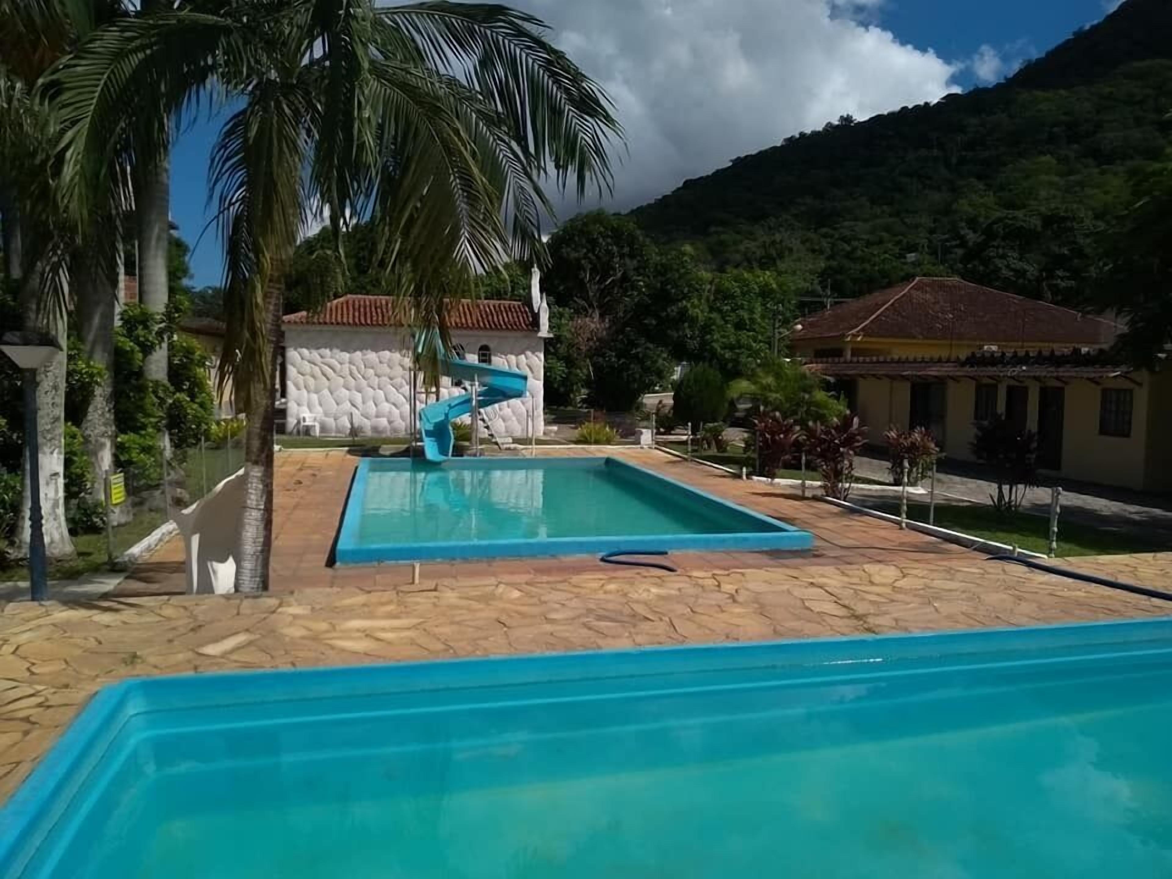 Piscina com escorregador, cercada por palmeiras e um cenário montanhoso.
