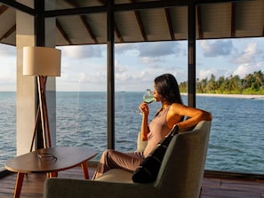 A person sitting by a window with a view of the sea, a table with a lamp, and a chair.
