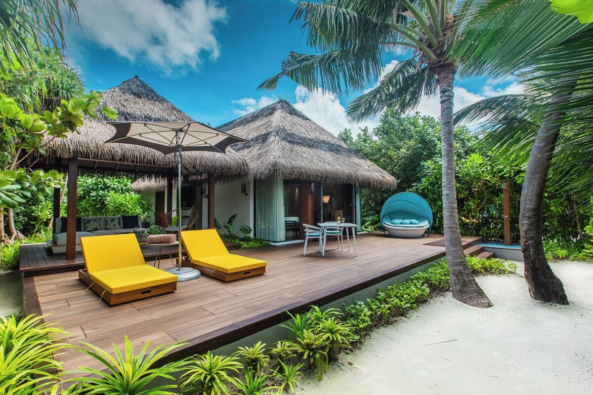 Un resort tropical con una terraza de madera, sillas reclinables amarillas, una estructura de techo de paja y una zona de piscina.