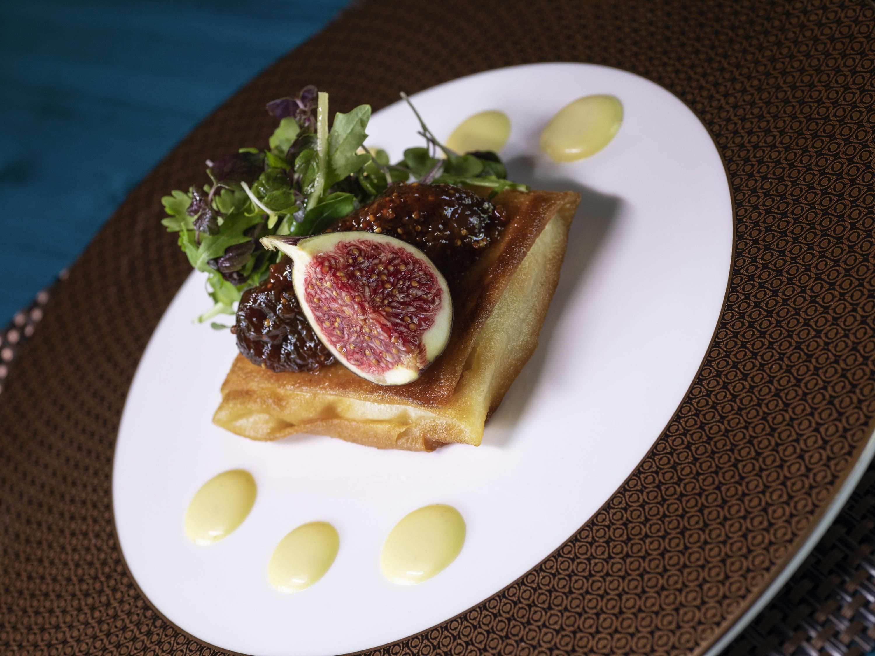 Une assiette carrée sur laquelle est disposé un aliment également de forme carrée, peut-être une tarte, garnie d’une tranche de figue et de quelques feuilles de verdure, le tout surmonté de trois cuillerées d’une sauce jaune.