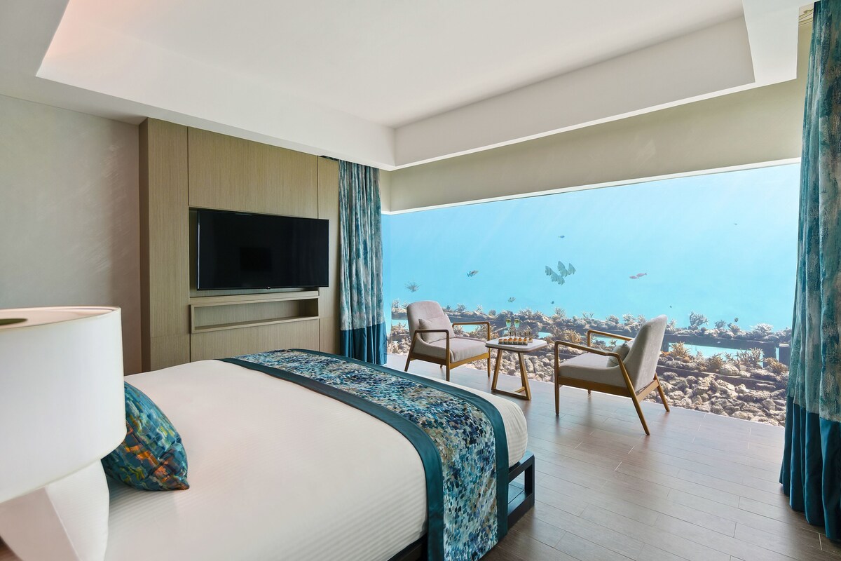 Habitación de hotel con cama, televisión, balcón con sillas y vista al océano.