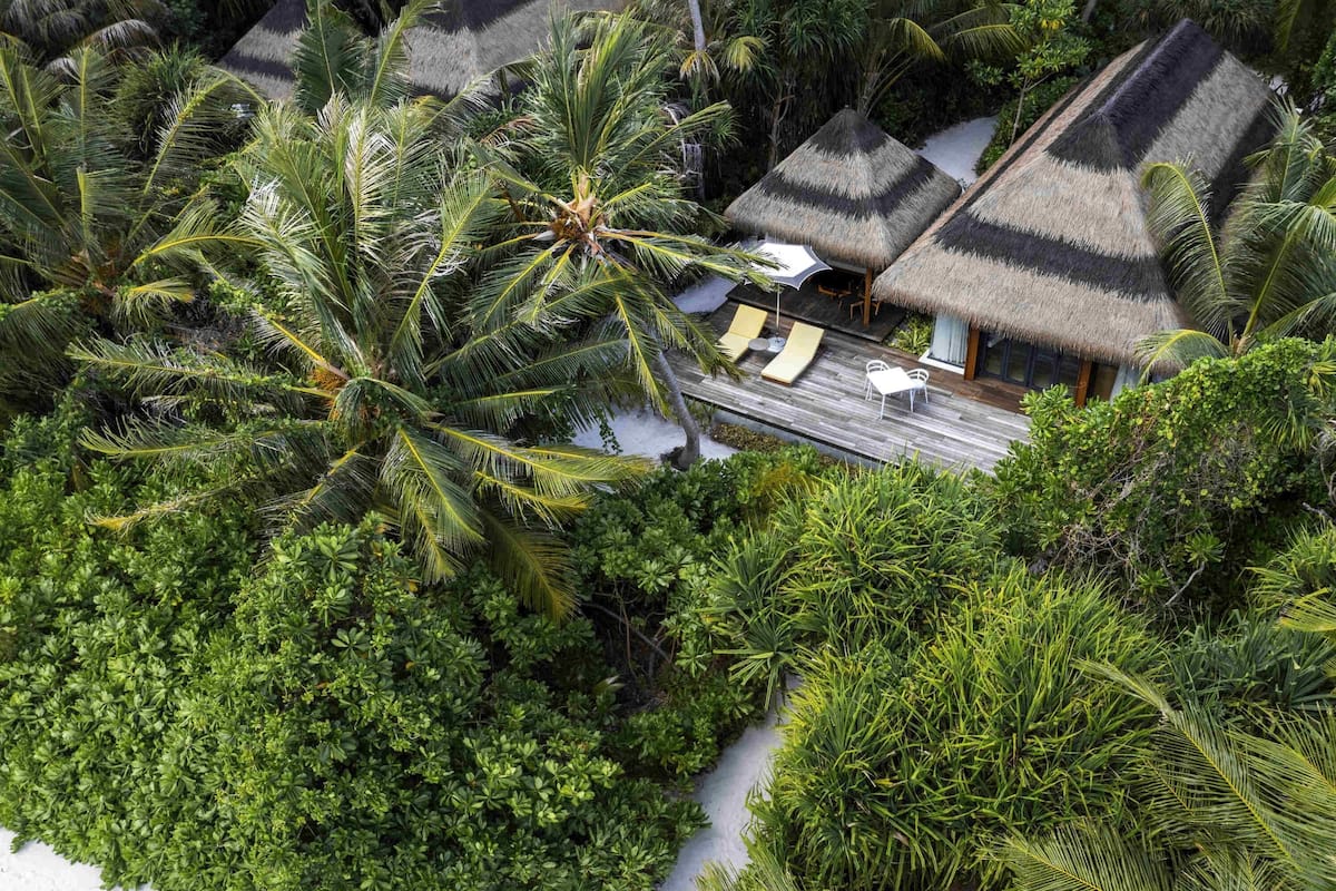 Vista aérea de un resort con estructuras de techo de paja, palmeras y una terraza de madera con sillas de descanso.