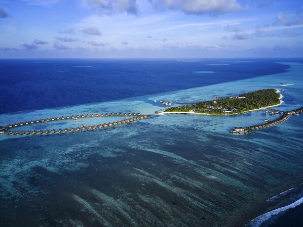 Eine Vogelperspektive einer Resorteinzelinsel mit überwasserbungalows, die in einer Sichelform angeordnet sind, umgeben von klarem, blauem Wasser und einem kleinen Festland mit Gebäuden.