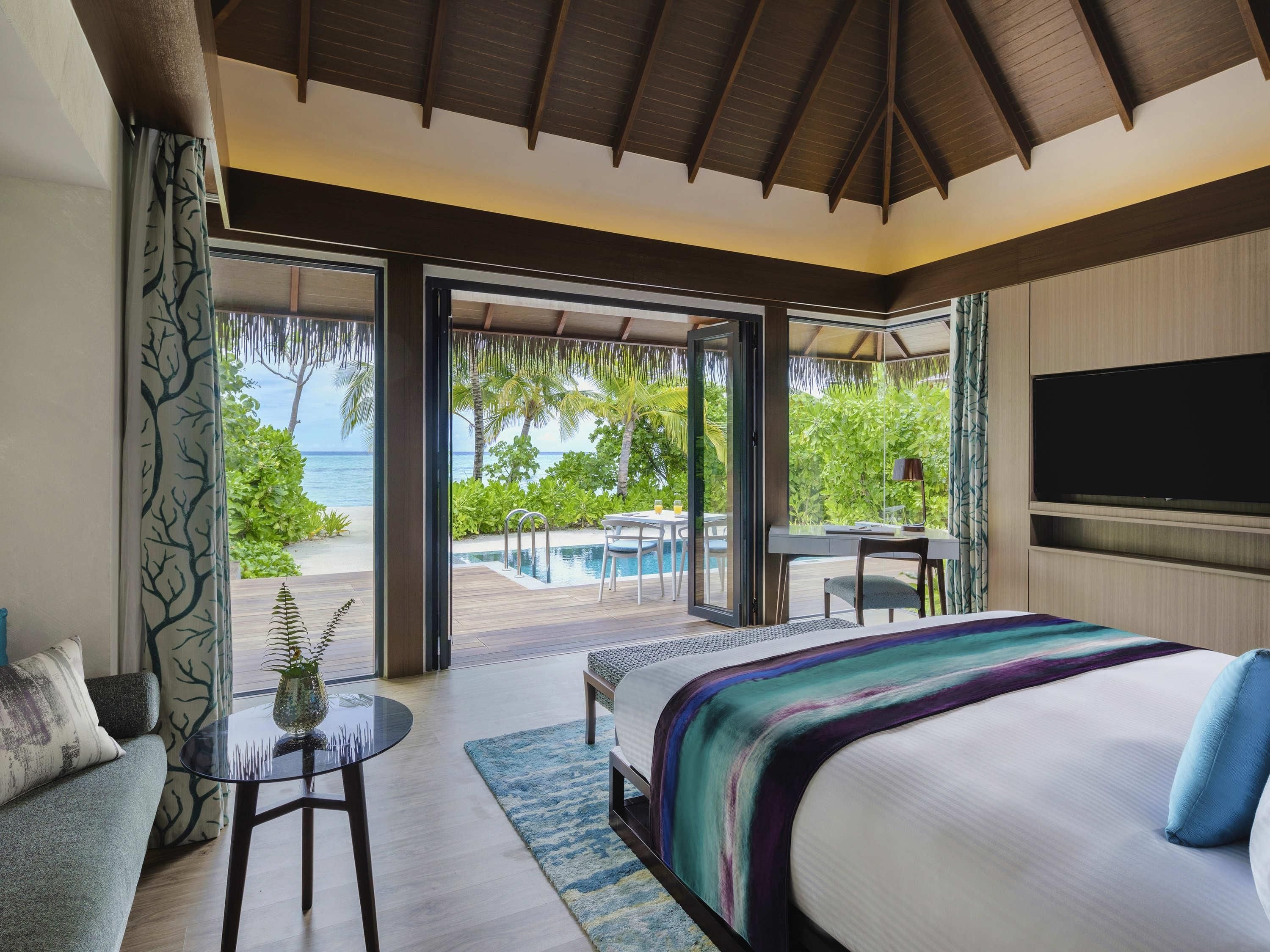 A bedroom with a large bed, a desk, and a TV. There is a view of the ocean and palm trees.