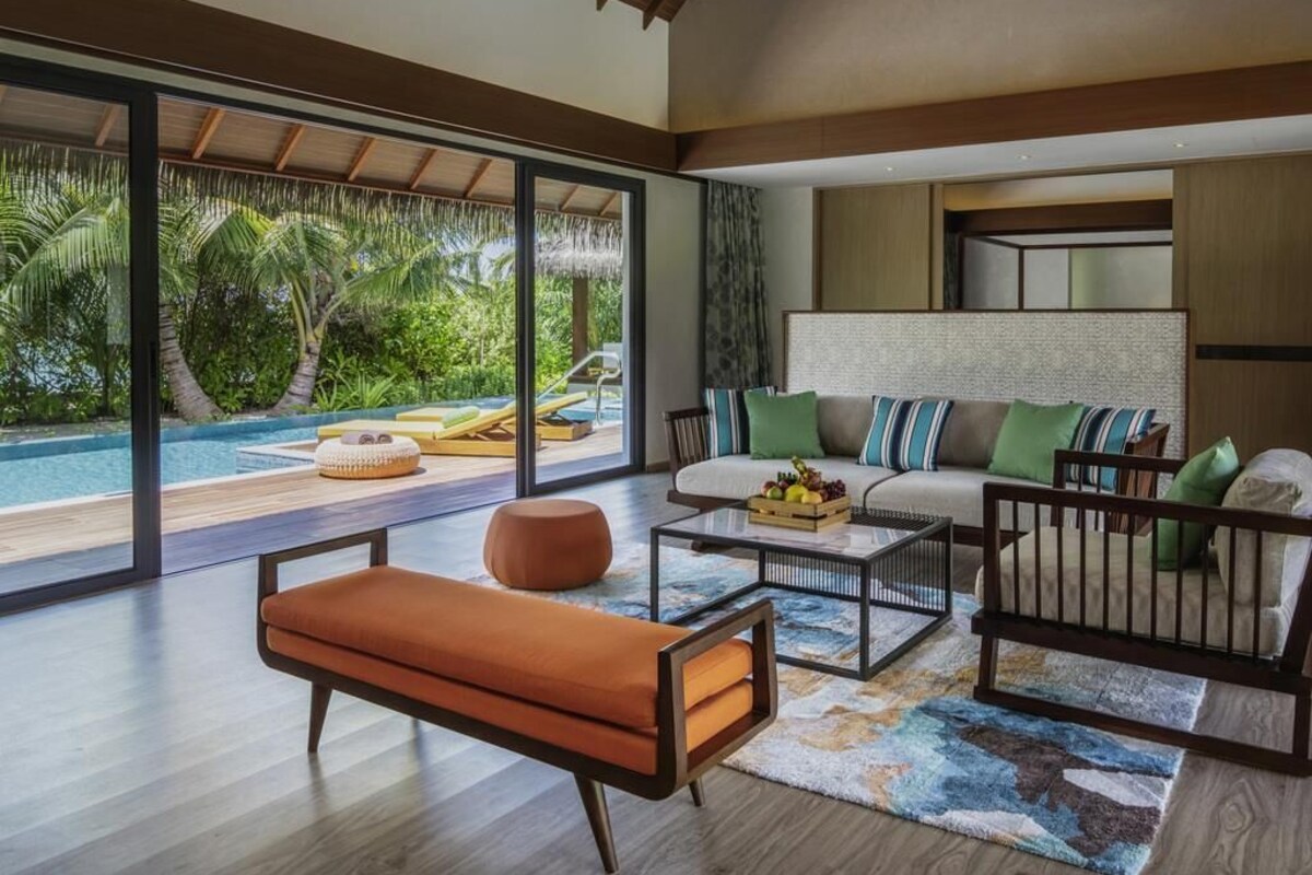 Una sala moderna con sofá, mesa de centro y reposapiés, un ventanal con vista a una piscina y follaje, y una puerta corrediza que da a otra habitación.