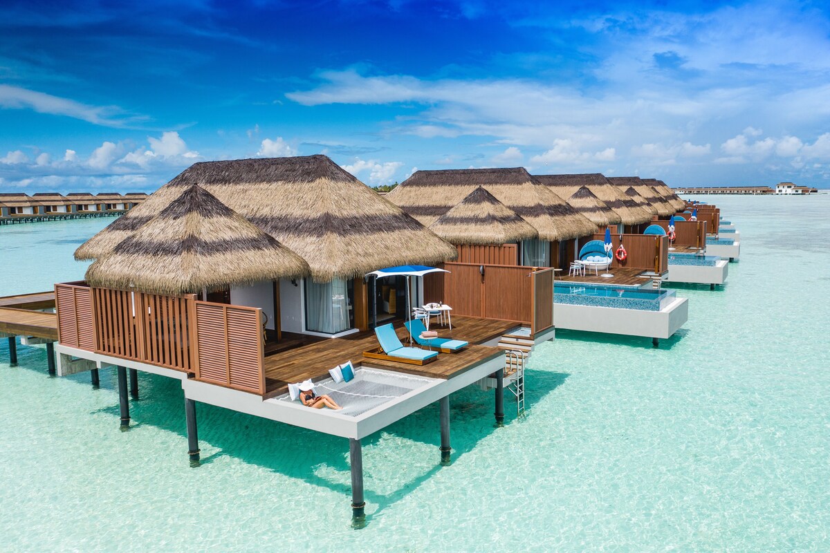 Bungalows sobre el agua con techos de paja, muebles de madera en terraza y una vista despejada del mar turquesa.