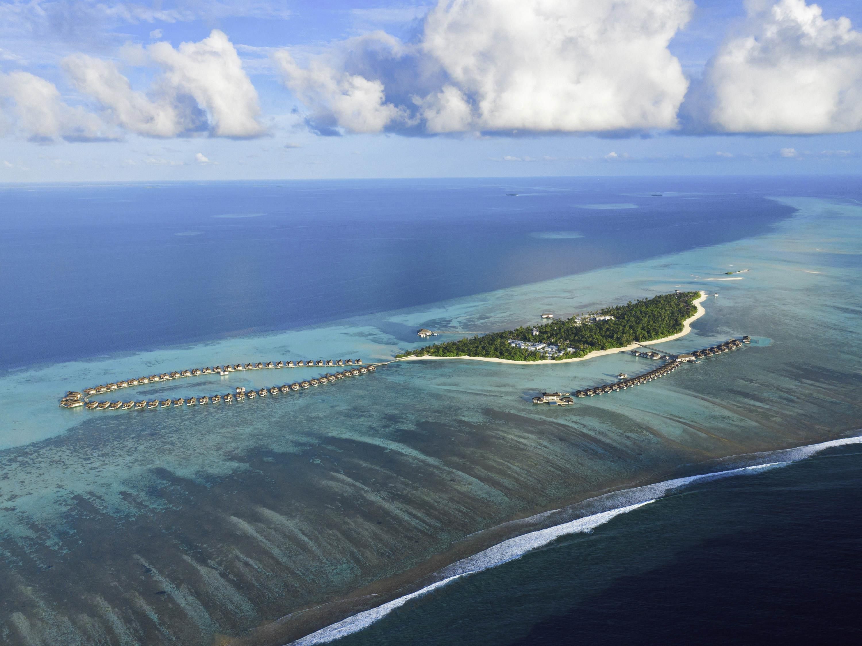 Luchtbeeld van een tropisch eilandresort met bungalows op palen, helder turquoise water en een zandstrand.