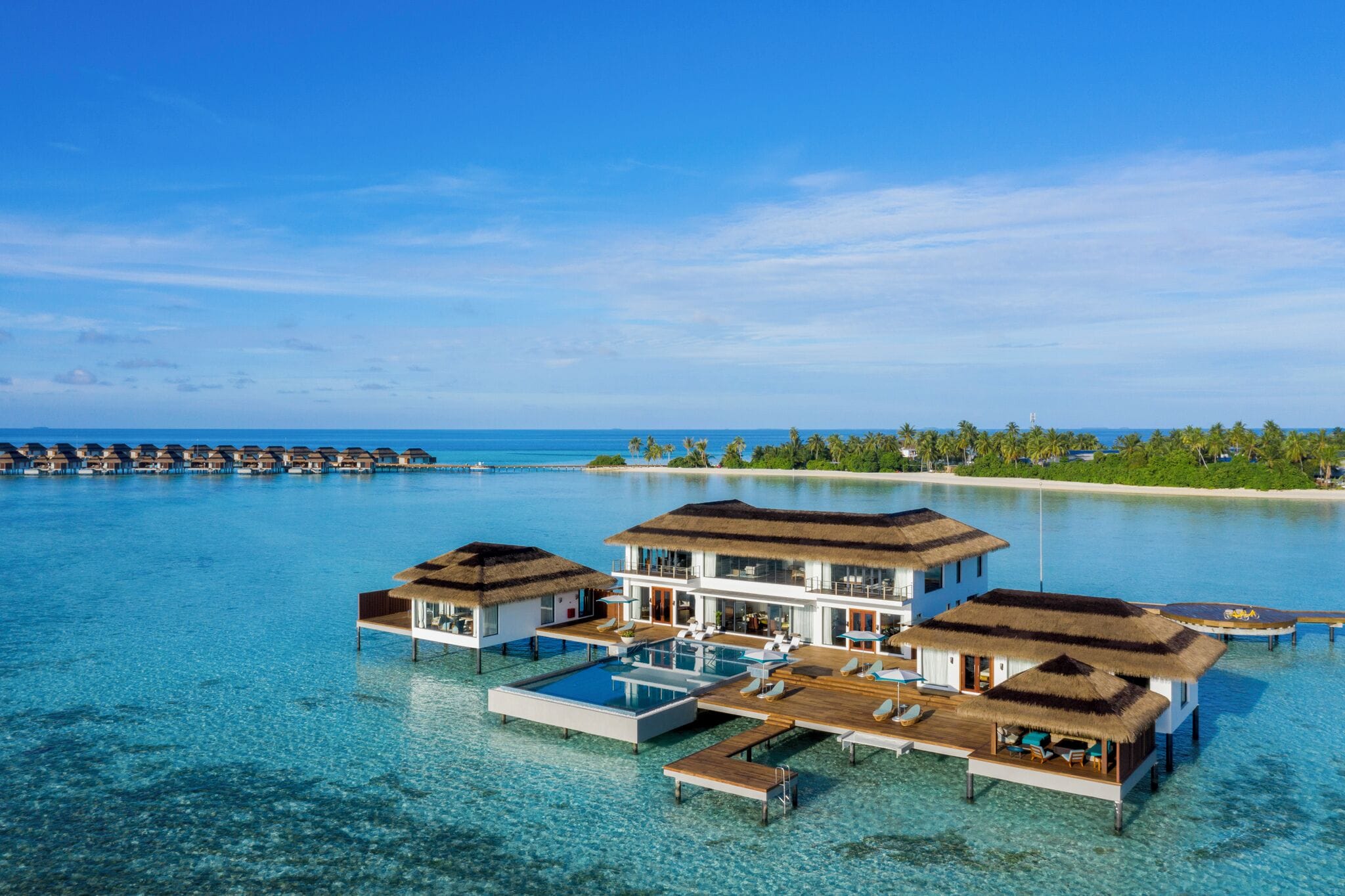 Overwater bungalows with infinity pools, a clear blue sea, and a tropical island in the background.