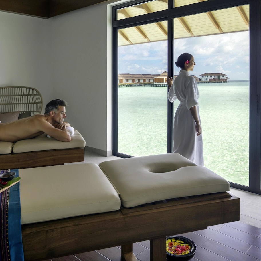 A room with two massage beds, a large window with a view of the ocean, and a small table with bowls of food.